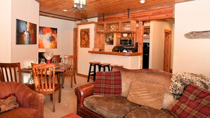 Una sala de estar acogedora con un sofá de cuero marrón, una mesa de comedor de madera y sillas. Se ve una cocina con armarios de madera y un microondas al fondo.