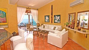 A living room with a white sofa, a coffee table, and a dining area with a table and chairs. There is a large window with curtains, a painting on the wall, and a shelf with decorative items.