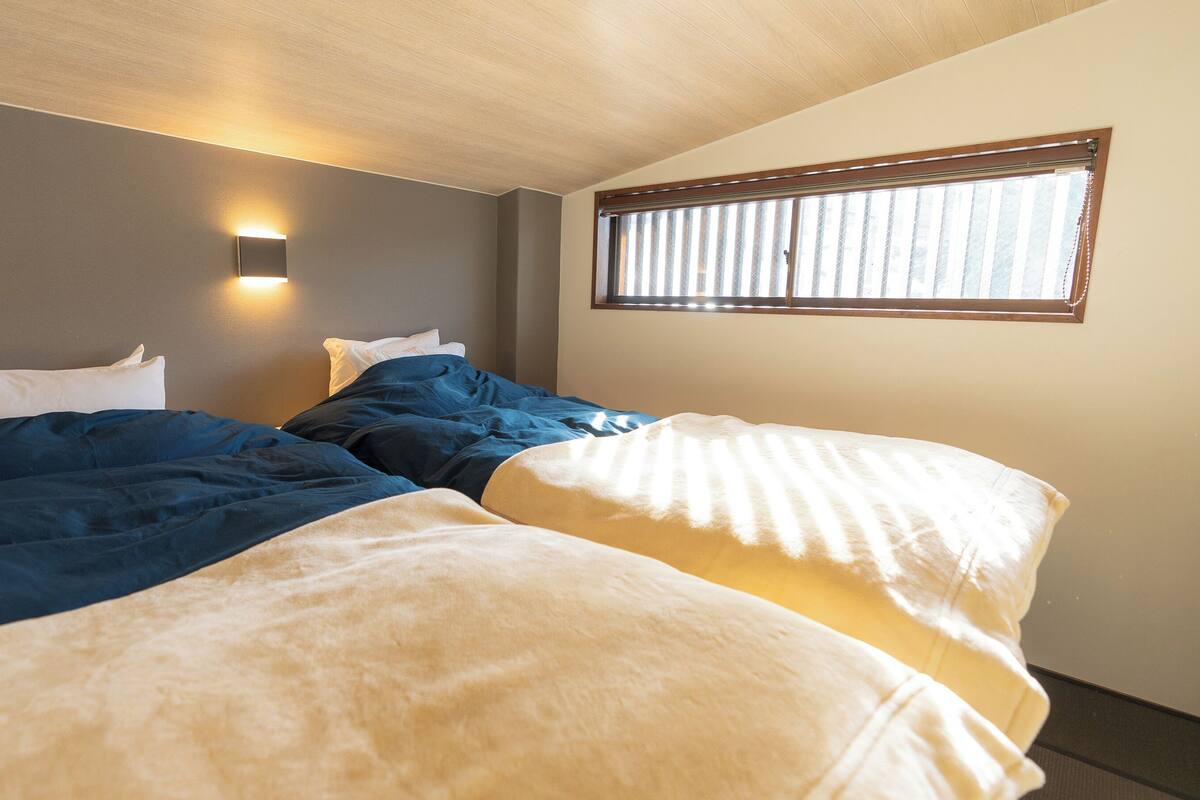 A hotel room with two beds, a wall-mounted light, and a window with blinds.
