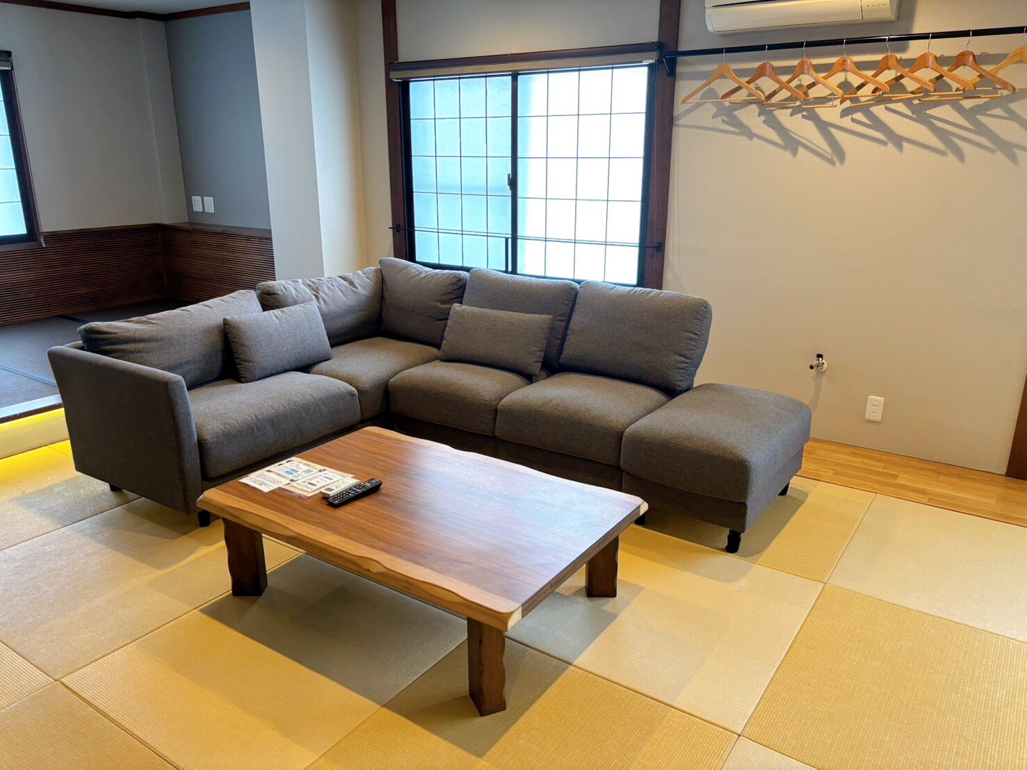 Una sala moderna con un sofá modular, una mesa de centro de madera, suelo de tatami, un ventanal grande y un aire acondicionado instalado en la pared.