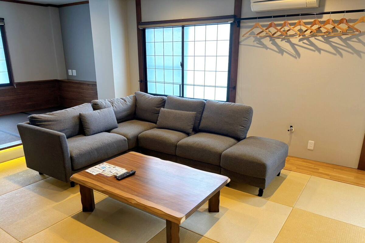 A modern living room with a sectional sofa, a wooden coffee table, tatami flooring, a large window, and a wall-mounted air conditioner.