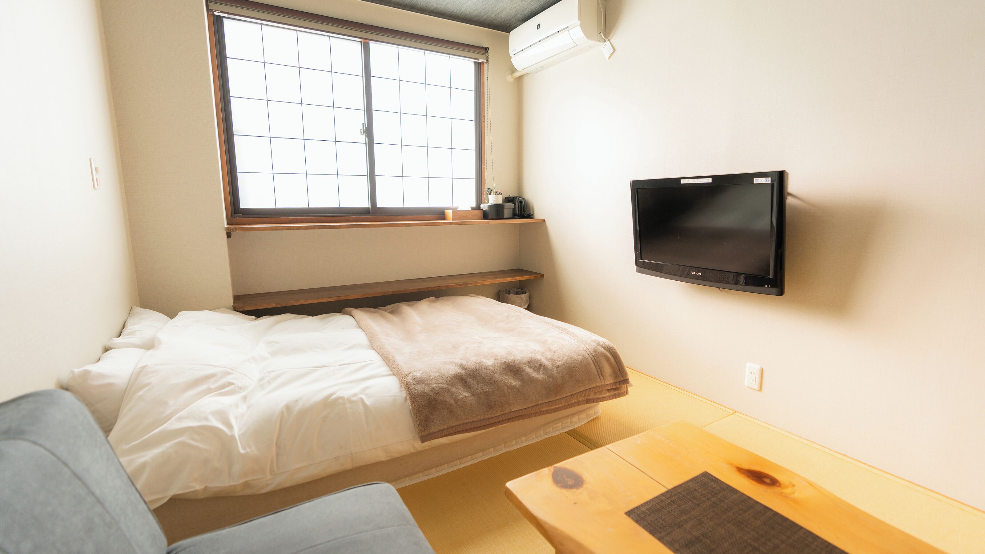 Una habitación pequeña y acogedora con una cama, un televisor montado en la pared, una ventana con persianas y un estante de madera sobre la cama.