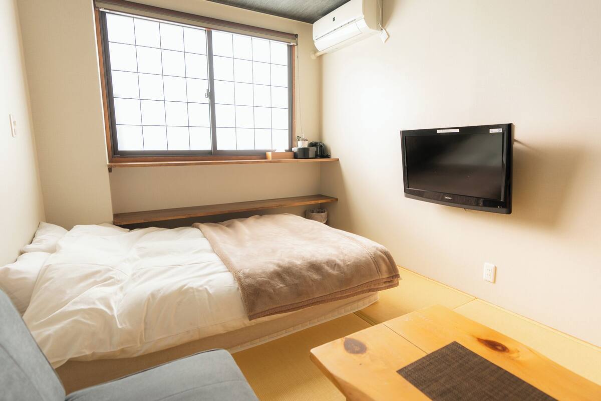 A small, compact room with a bed, a TV mounted on the wall, a window with blinds, and a wooden shelf above the bed.