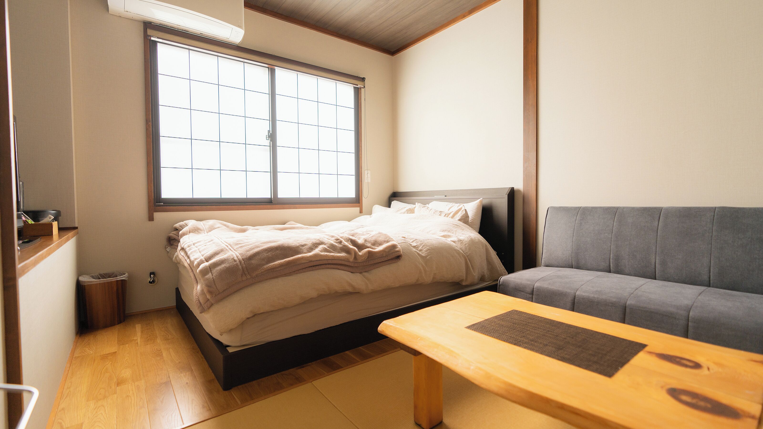 Un dormitorio con una cama, un sofá gris, una mesa de madera y una ventana con persianas.