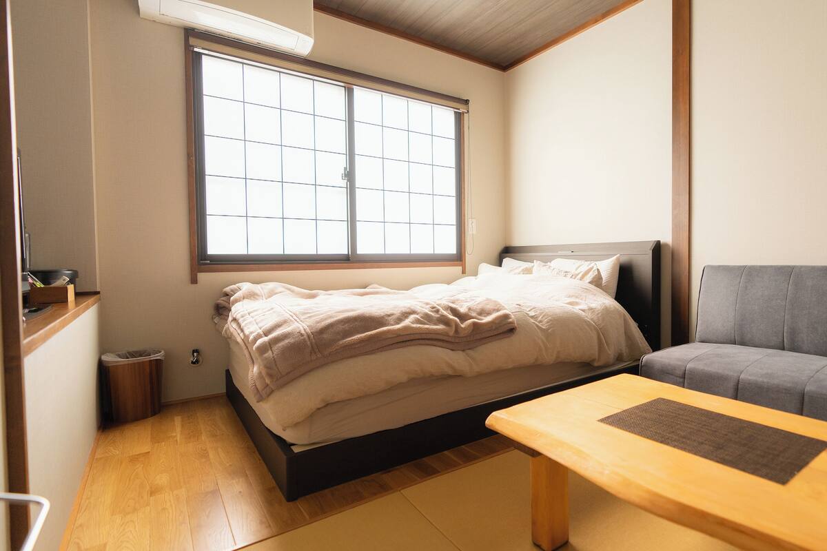 A bedroom with a bed, a grey sofa, a wooden table, and a window with blinds.