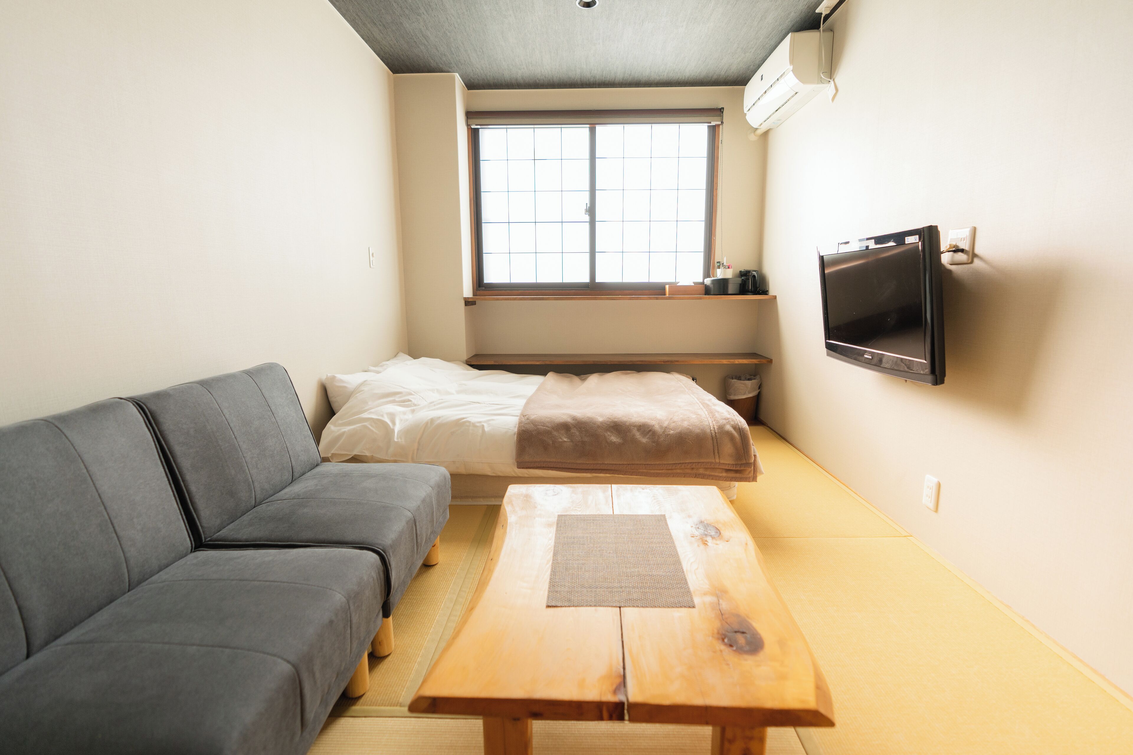 A compact room with a sofa, a bed, a wooden table, and a TV mounted on the wall.