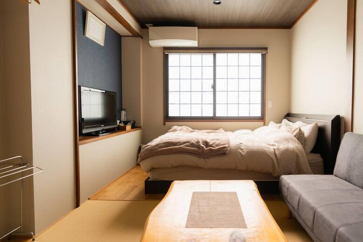 A compact room with a bed, a gray sofa, a TV, and a window.