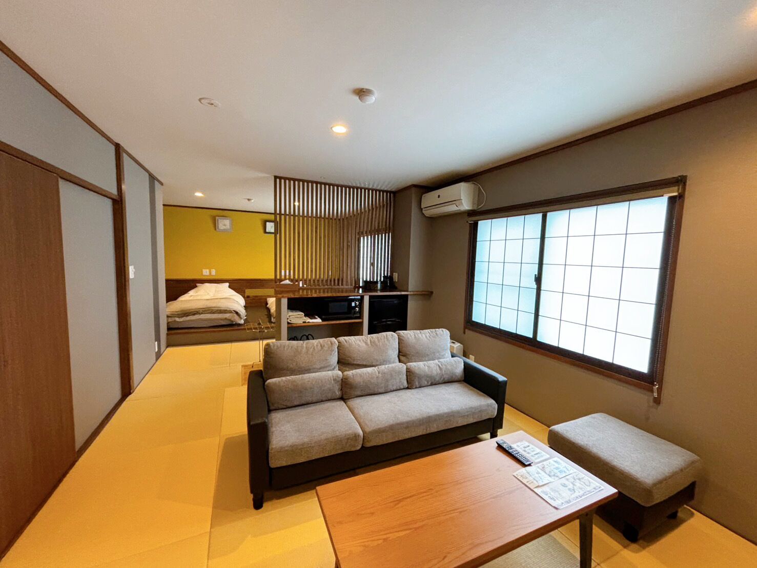 Una habitación moderna con sofá, una mesita, una banca, ventana con persianas, televisor y zona de kitchenette.
