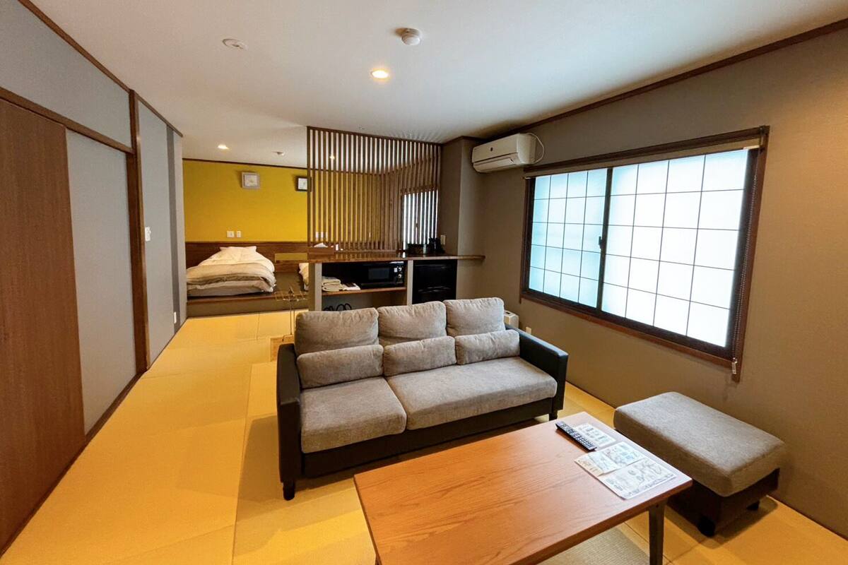 A modern room with a sofa, a small table, a bench, a window with blinds, a TV, and a kitchenette area.