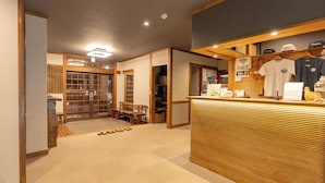 Una zona d'entrada amb un taulell de fusta, una zona de seients amb una banca, i vista a una sala tradicional japonesa amb portes corredisses.