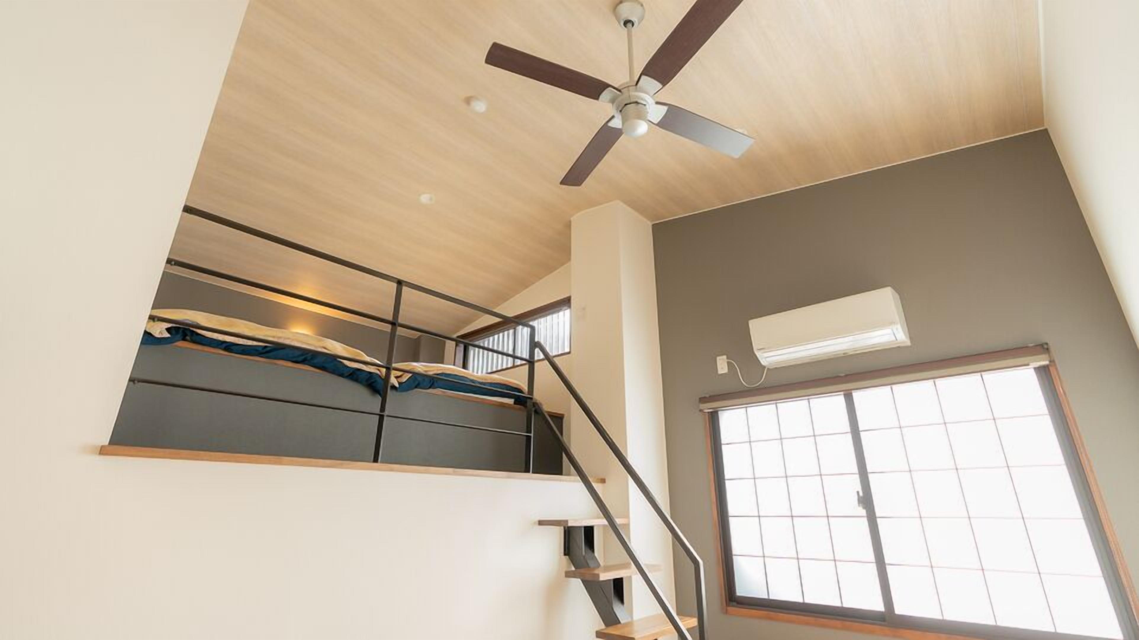Una habitación con ventilador de techo, ventana con persianas, una escalera y un equipo de aire acondicionado.