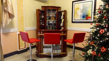 A corner bar area with three red bar stools, a wooden bar cabinet with glassware, a framed abstract painting, and a decorated Christmas tree.
