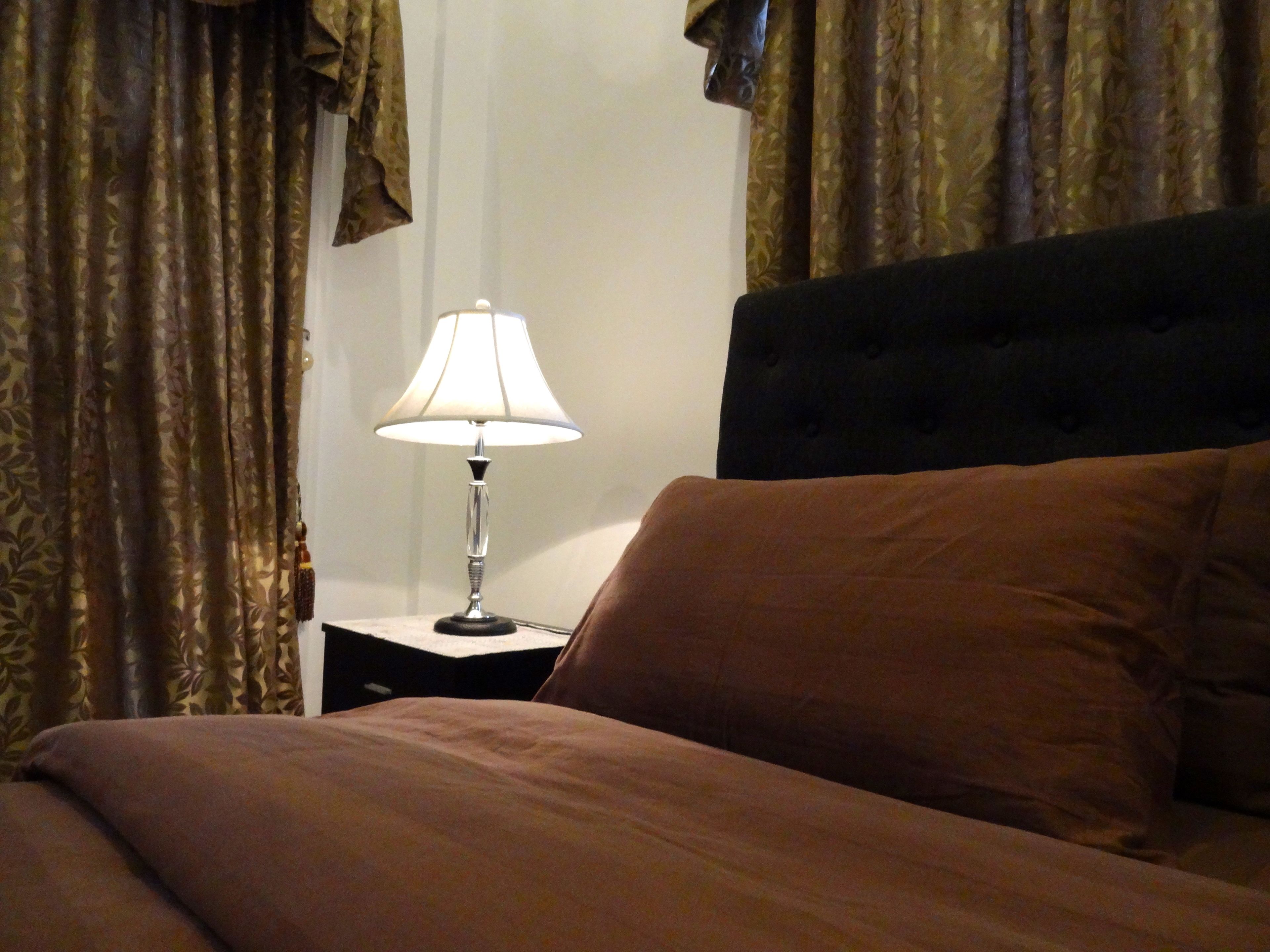A neatly made bed with a brown comforter and pillows, a bedside lamp on a nightstand, and curtains with a leaf pattern.