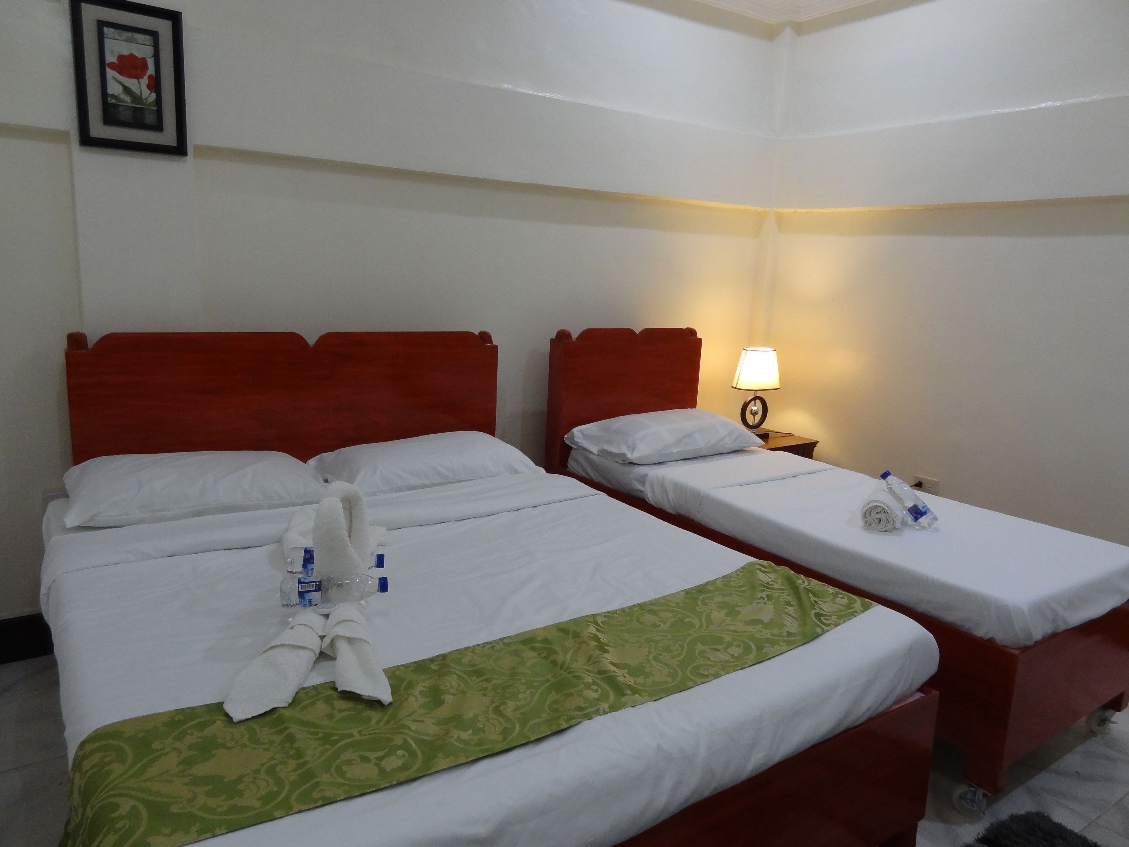 Habitación de hotel con dos camas, cada una con ropa de cama blanca y cabeceros rojos. Un corredor de cama verde con estampado está sobre la cama inferior. Se ve una mesita de noche con una lámpara y una botella de agua.