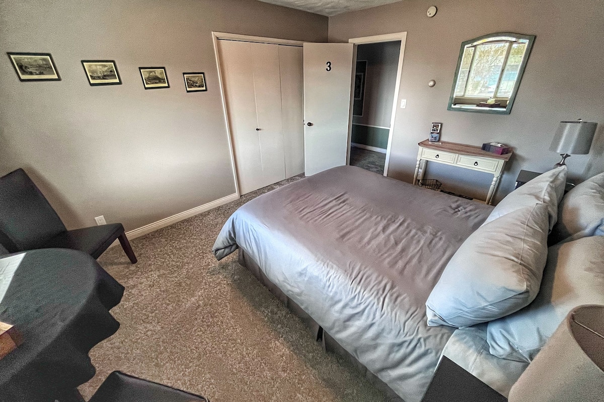 Un dormitorio con cama, escritorio, silla, armario y una mesita de noche con lámpara.