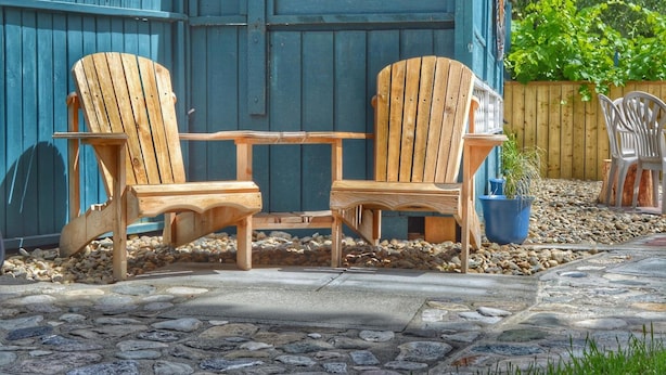 Duas cadeiras de madeira estilo Adirondack com uma mesinha no meio, dispostas num pátio de pedra e com um muro de madeira ao fundo.