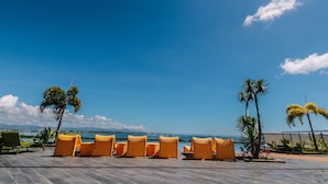 A poolside area with lounge chairs and palm trees, overlooking a body of water and mountains. - CSuites at 8 Newtown Residences (Lapu-Lapu)