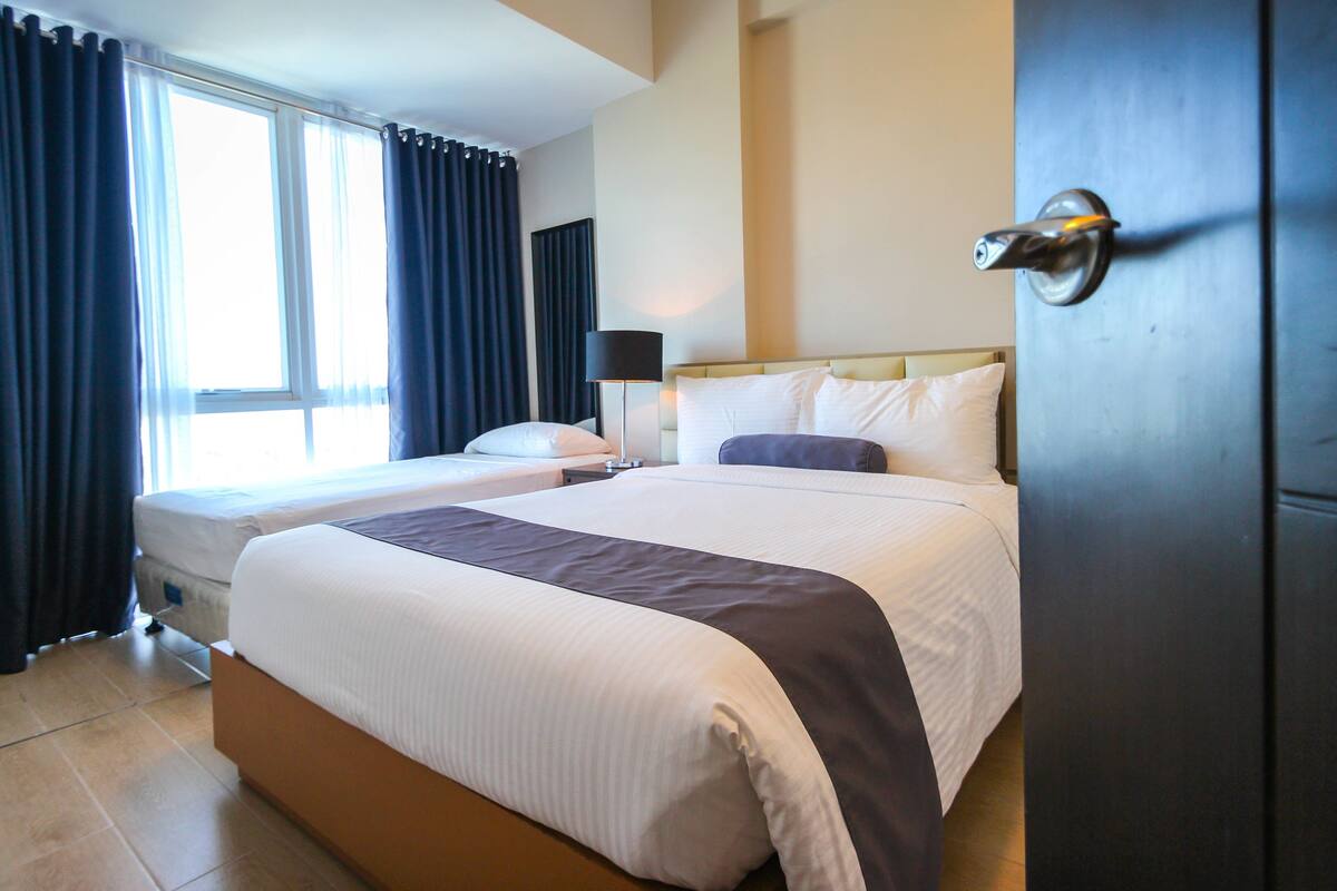 Une chambre d’hôtel avec deux lits, une grande fenêtre avec des rideaux, une lampe de chevet et une porte.