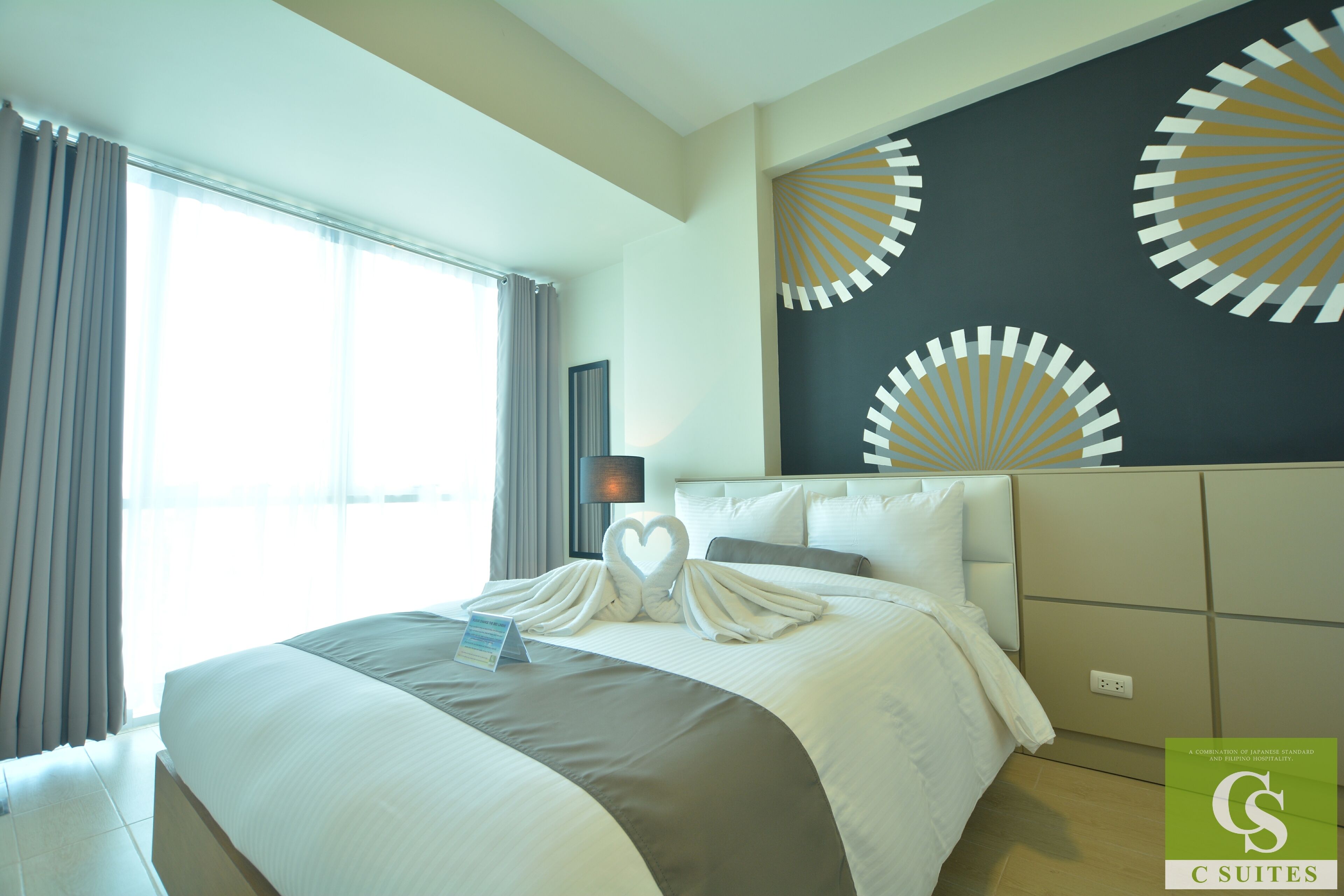 A hotel room with a large bed, a headboard with circular patterns, a bedside table with a lamp, and a window with curtains.