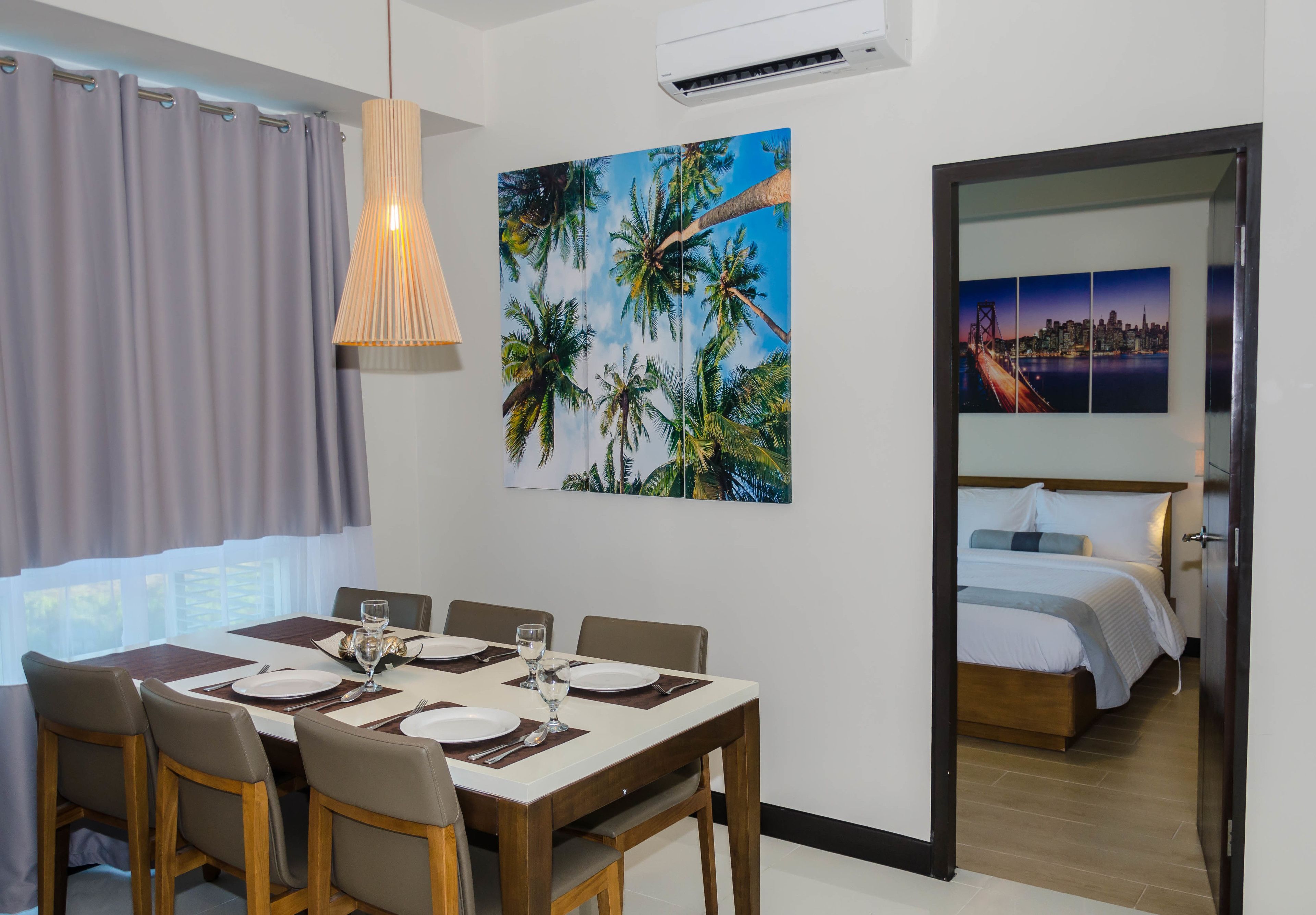 Uma sala de jantar com mesa posta para quatro pessoas, um quadro de palmeiras e um quarto visível por uma porta aberta.