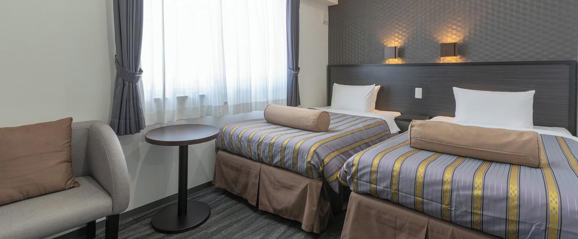 Habitación de hotel con dos camas, una mesita redonda pequeña, un sofá y una ventana con cortinas.