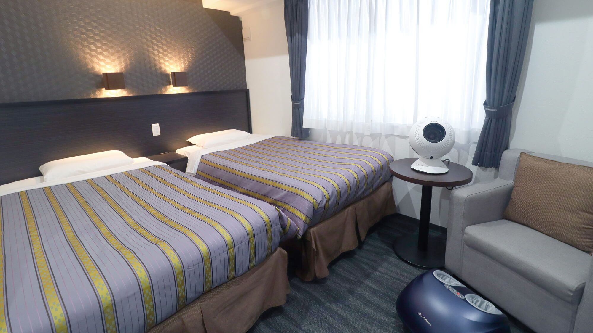 Habitación de hotel con dos camas, una mesita con una webcam, una silla y un sillón de masaje.