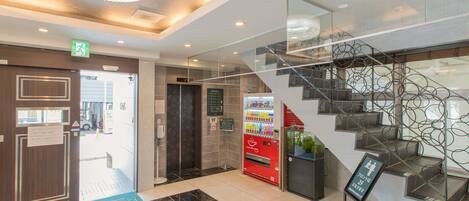 A modern hotel lobby with a marble floor, a staircase with a unique metal design, a vending machine, and a digital elevator display.