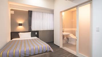 Ein Hotelzimmer mit Bett, Nachttisch, einer an der Wand befestigten Lampe, einem Badezimmer mit Waschbecken und Toilette sowie einem Schiebetürenschrank.