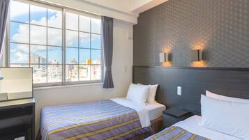 Quarto de hotel com duas camas, uma janela grande com cortinas, uma escrivaninha com abajur e vista para a cidade.