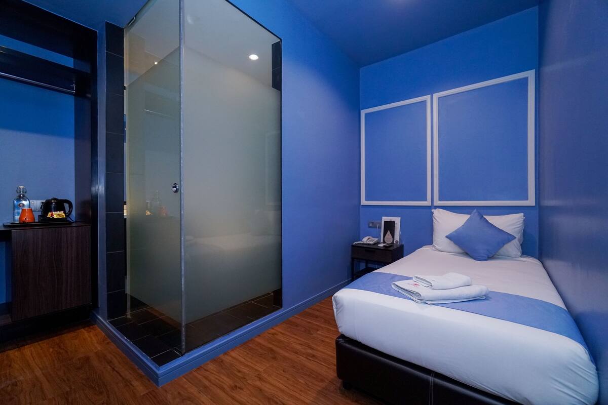 A modern hotel room with a glass-enclosed shower, a neatly made bed with white linens, and a nightstand with a phone and a lamp.