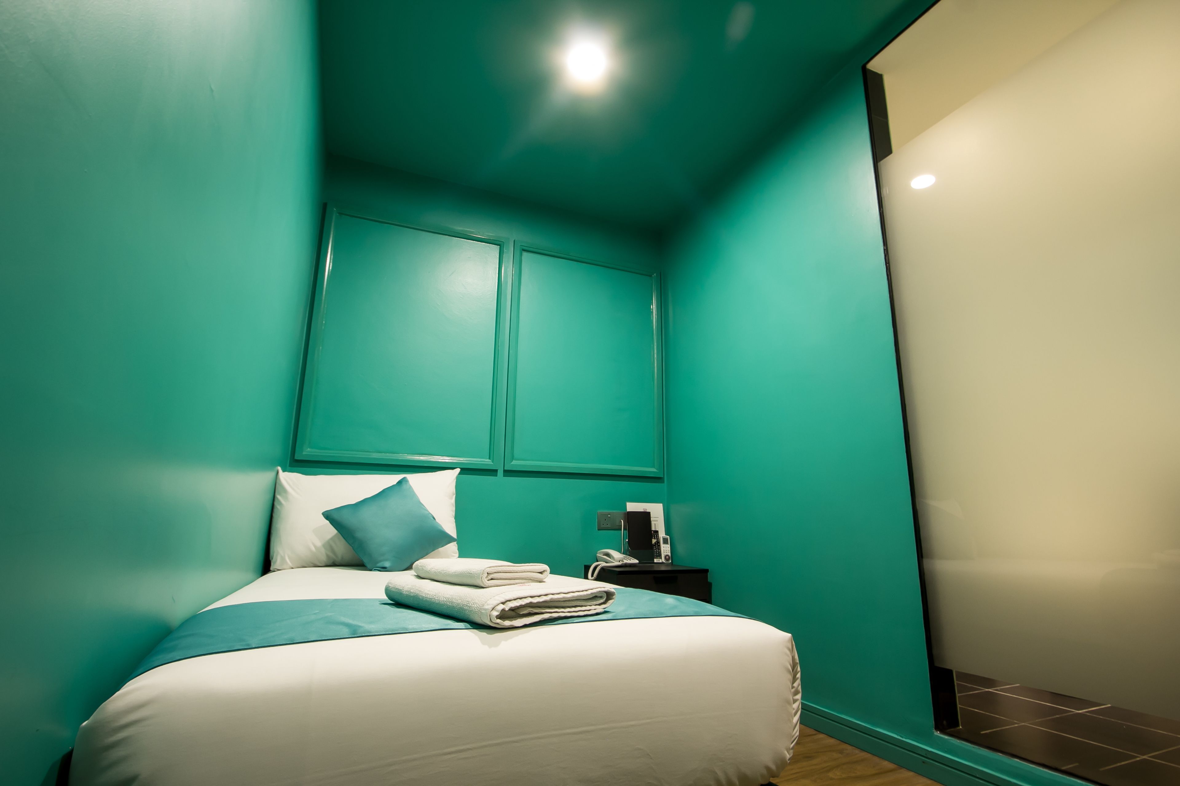 A neatly made bed with pillows and folded towels, a bedside table with a phone and a small device, and teal walls with a window on the right side.