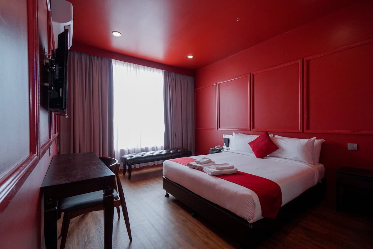A hotel room with a large bed, a desk, and a chair. There is a window with curtains, a television mounted on the wall, and a red accent wall.