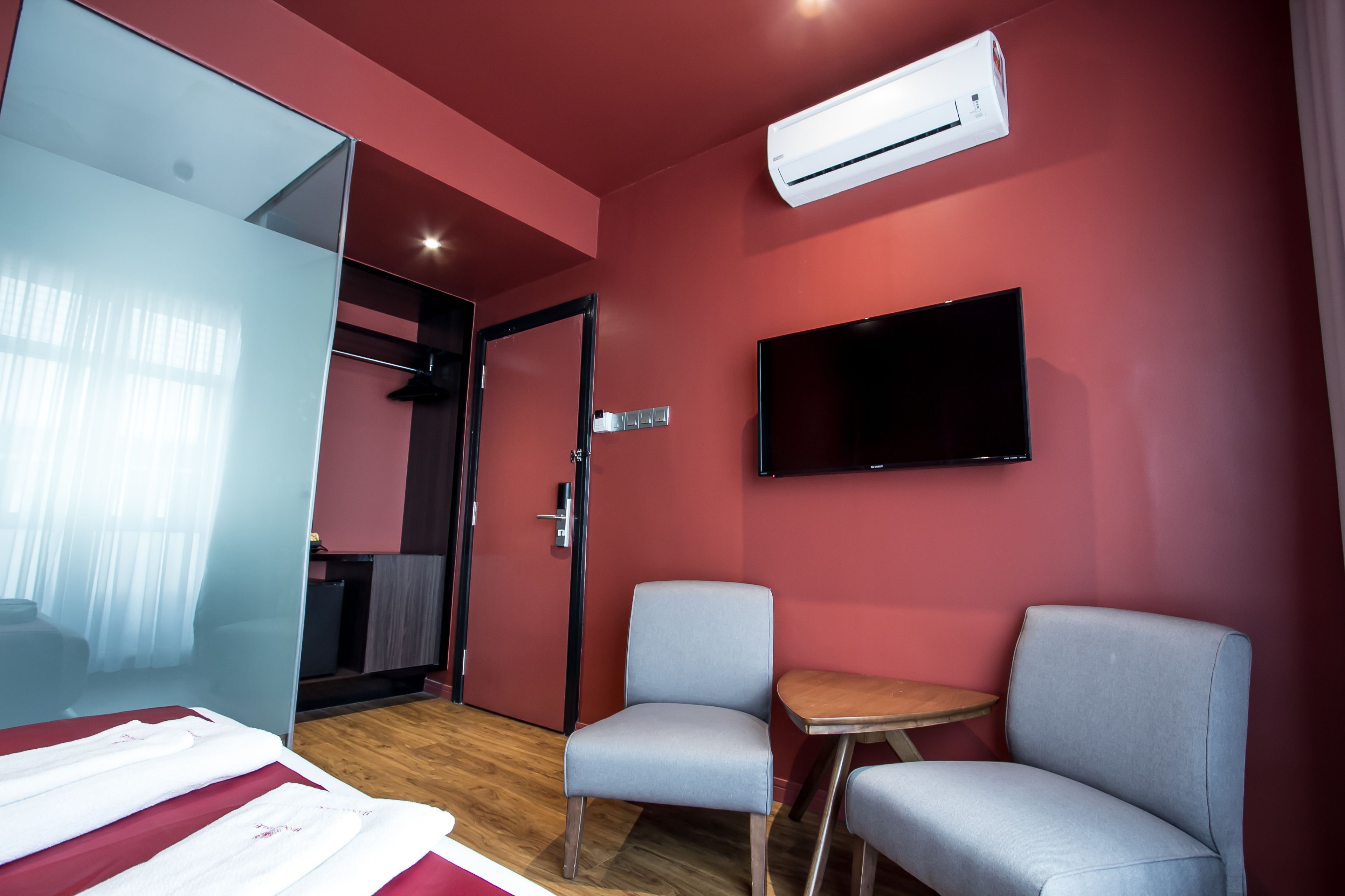 A hotel room with a bed, two grey armchairs, a small wooden table, a flat-screen TV mounted on the wall, and an air conditioning unit.