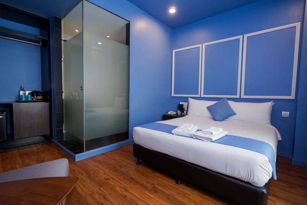 A modern hotel room with a large bed, a glass shower enclosure, a small desk with toiletries, and a blue accent wall.