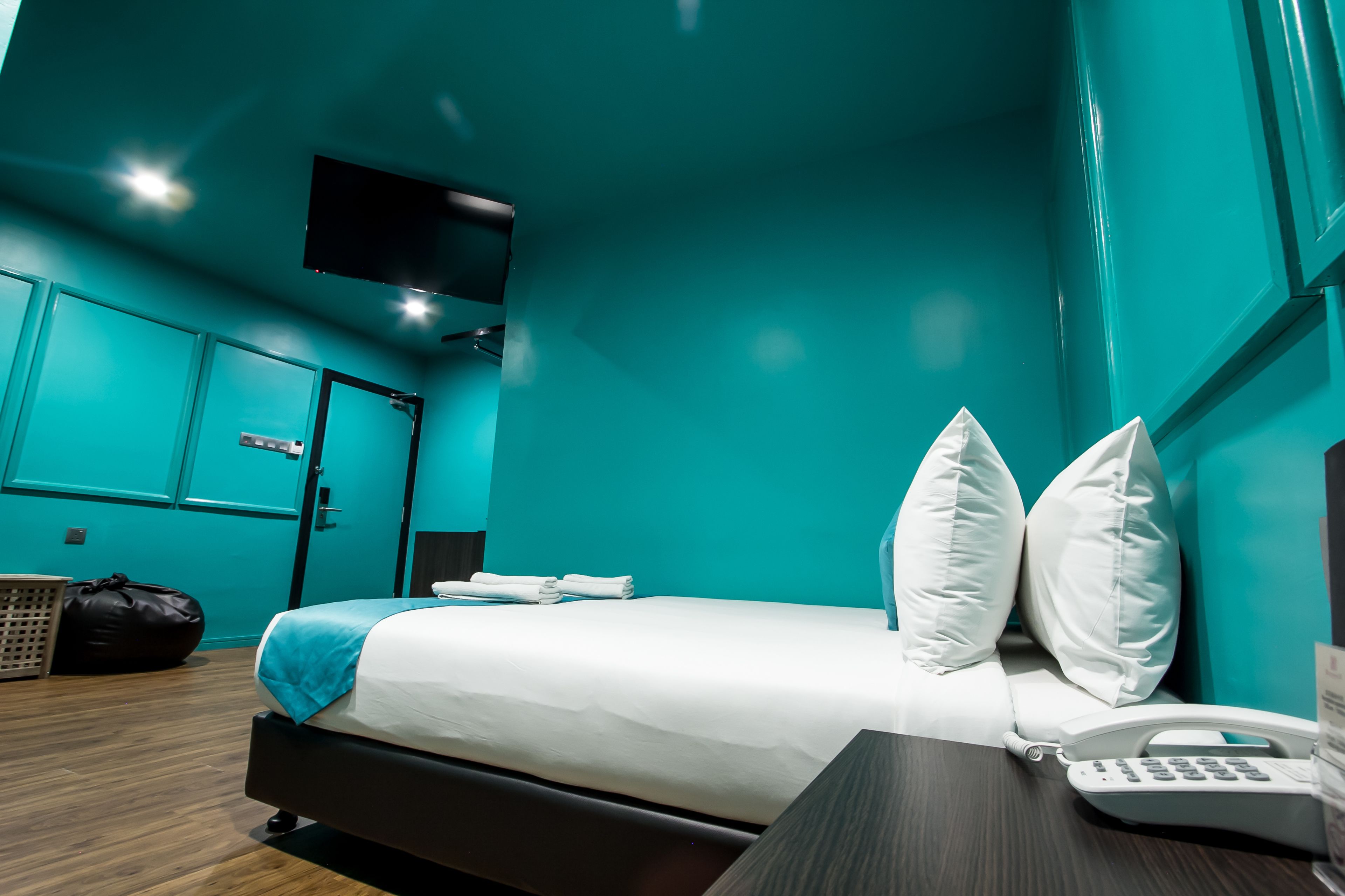 Una habitación de hotel moderna con paredes verde azulado, una cama con ropa de cama blanca, un escritorio con teléfono y un televisor montado en la pared.