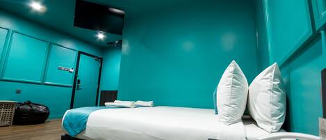 Una habitación de hotel moderna con paredes verde azulado, una cama con ropa de cama blanca, un escritorio con teléfono y un televisor montado en la pared.