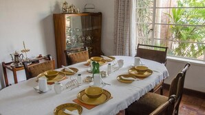 Una mesa de comedor dispuesta con vajilla dorada y blanca, un aparador de madera con puertas de vidrio y una ventana con follaje en el exterior.
