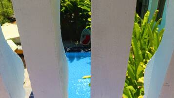 Vista a través de una cerca blanca, con una piscina azul y plantas verdes al fondo.