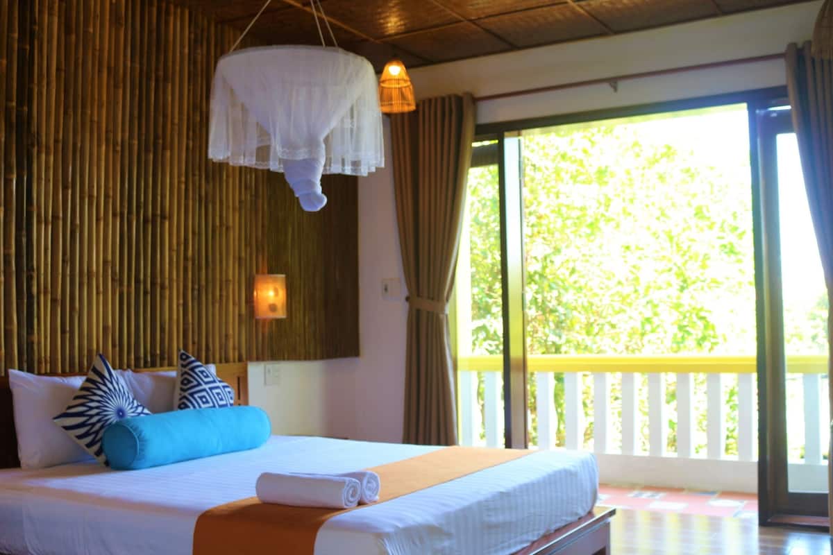 A room with a bed, a hanging canopy, a window with a view of greenery, and a balcony with a railing.