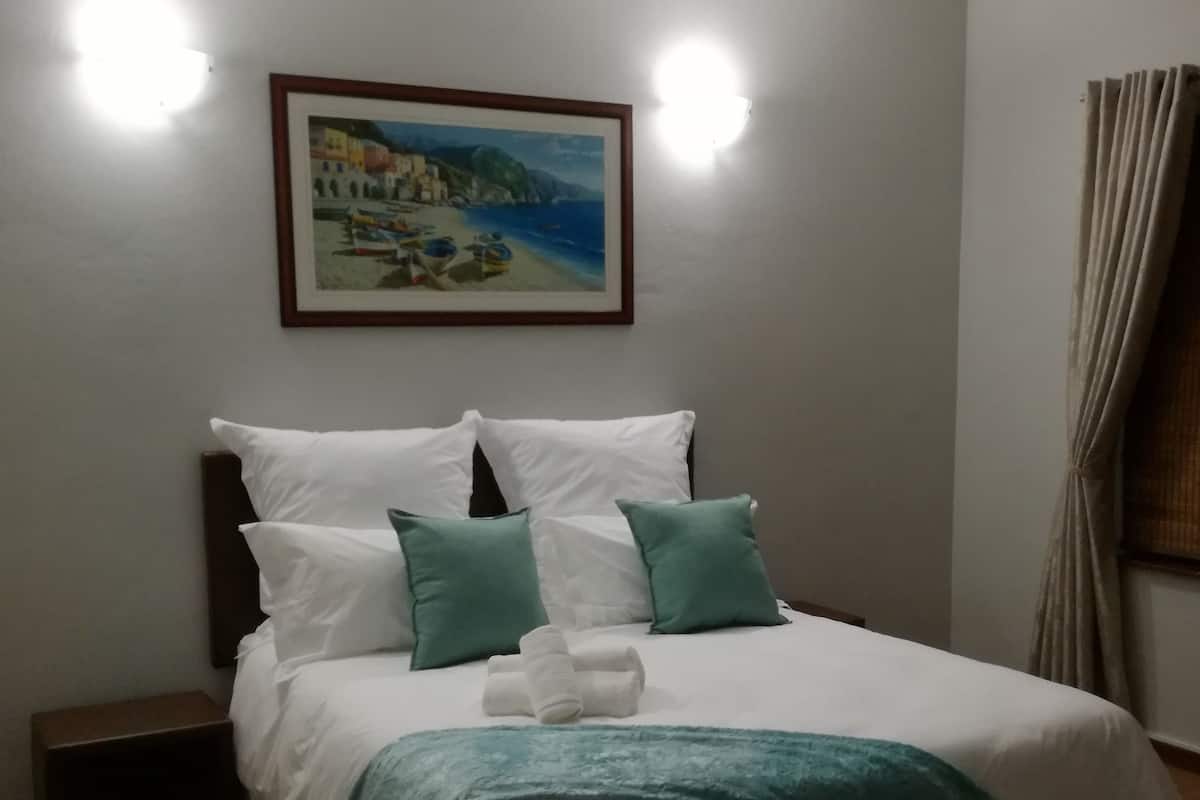 Ein ordentlich bezogenes Bett mit weißer und türkisfarbener Bettwäsche, ein kleiner Nachttisch, ein gerahmtes Bild an der Wand und ein Deckenventilator.