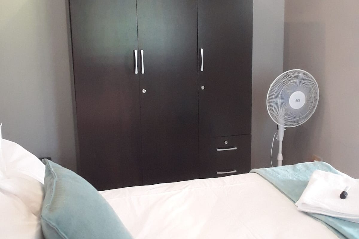 Ein Schlafzimmer mit Bett, Kissen, einem Kleiderschrank und einem Standventilator.