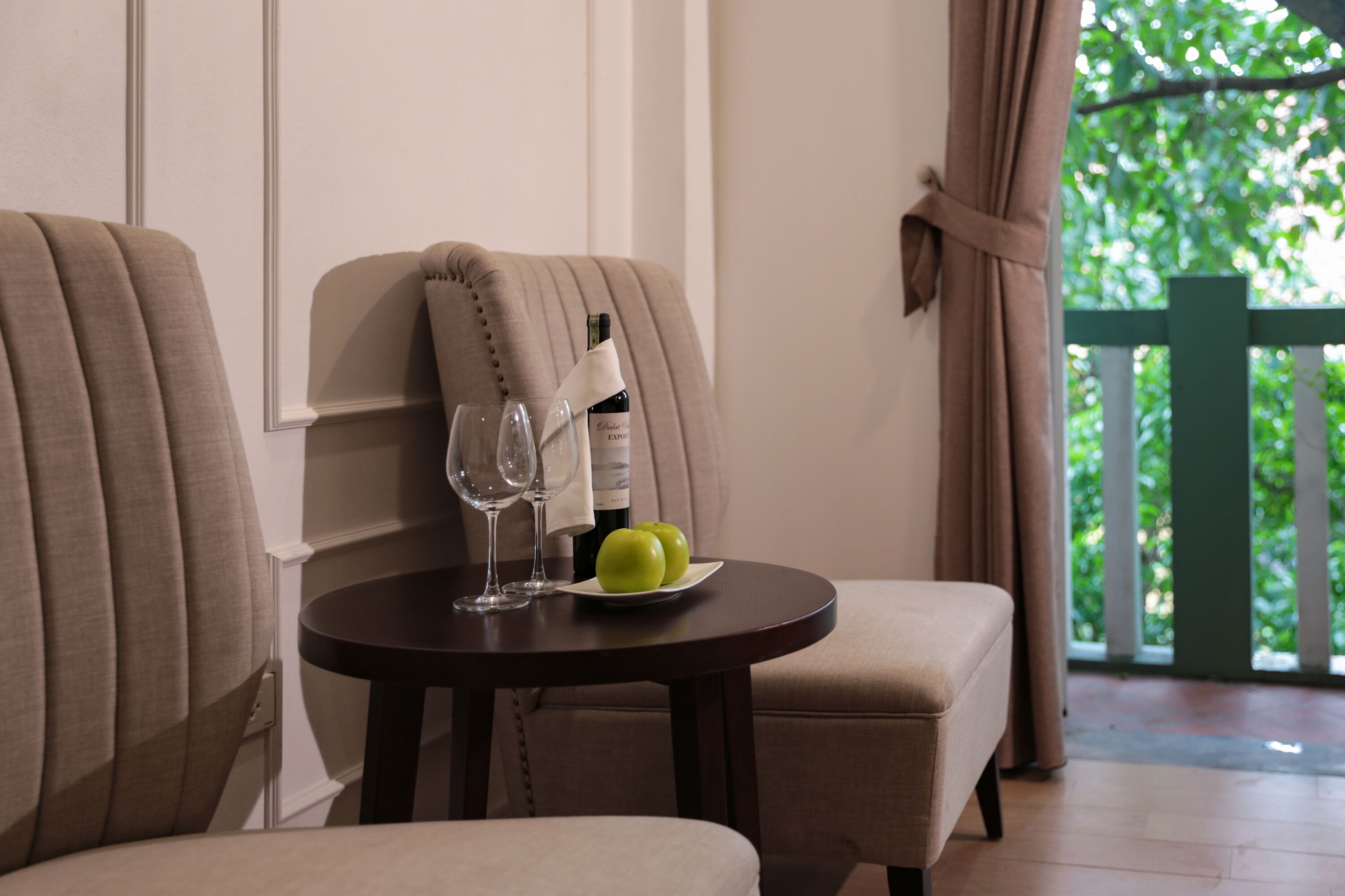 A dining area with two upholstered chairs, a round table, a bottle of wine, two glasses, and a plate with two green apples.
