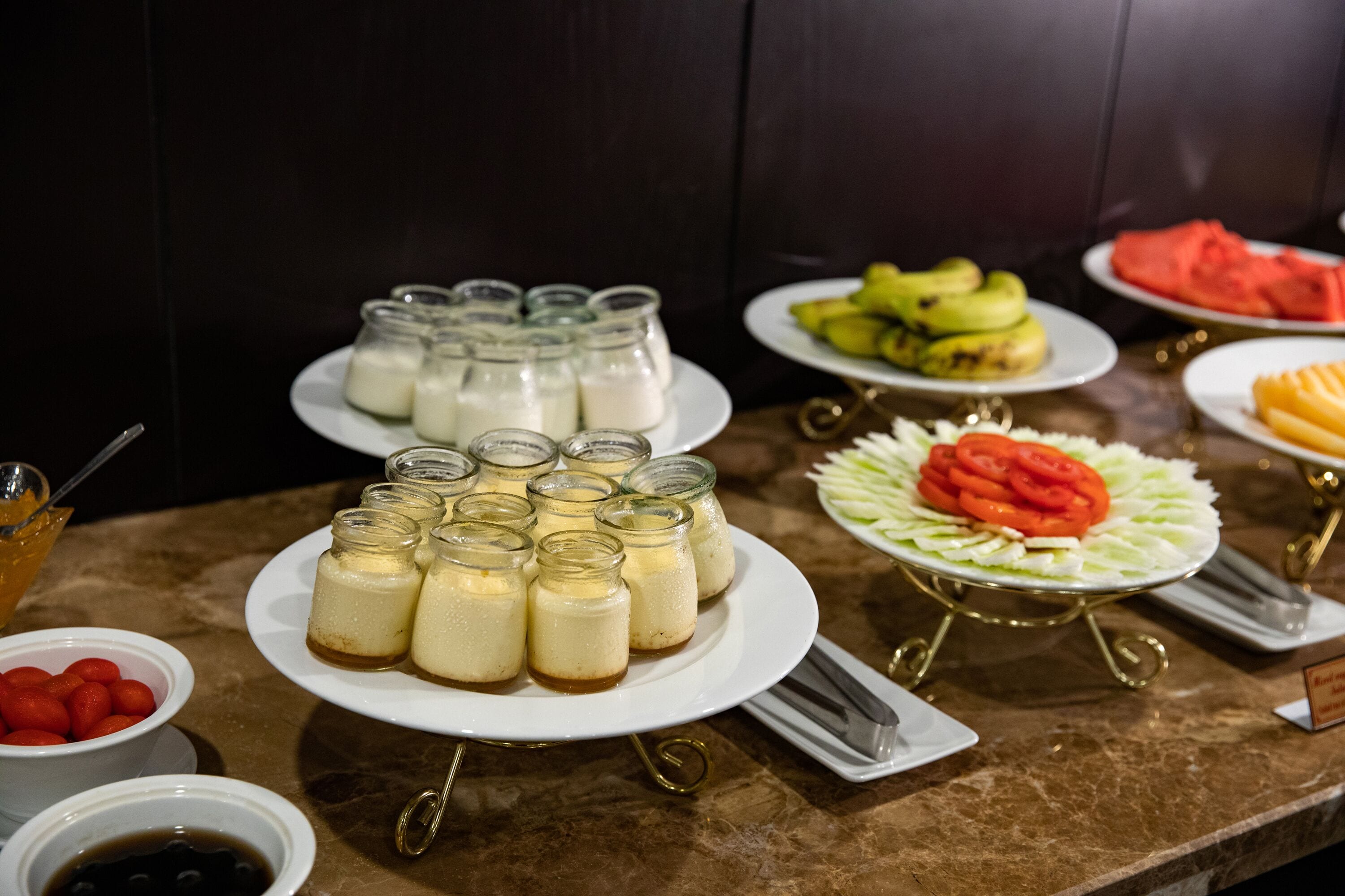 Una mesa de buffet con diferentes opciones de comida, incluyendo botes de un postre cremoso, frutas en rodajas y un plato con una presentación decorativa de tomates y lechuga.