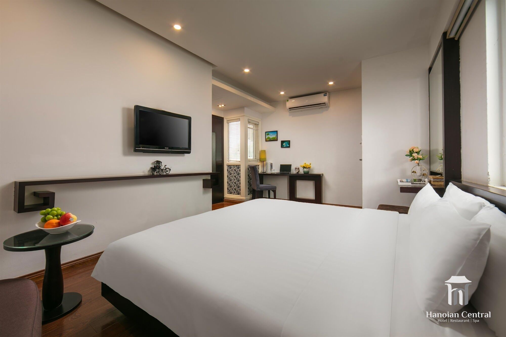 A hotel room with a large bed, a TV mounted on the wall, a small desk with a chair, and a window with curtains.