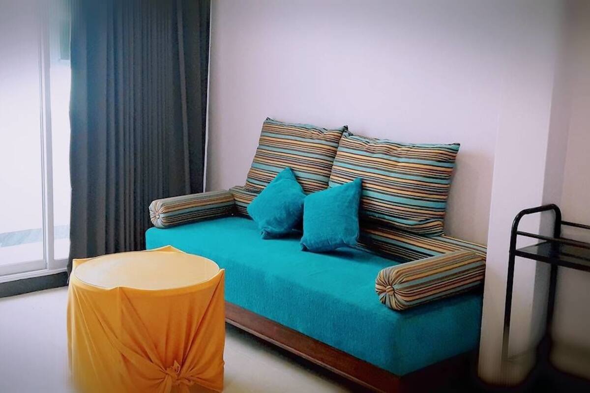 Ein Zimmer mit einem blaugrünen Sofa, gestreiften Kissen und einem gelben Hocker.