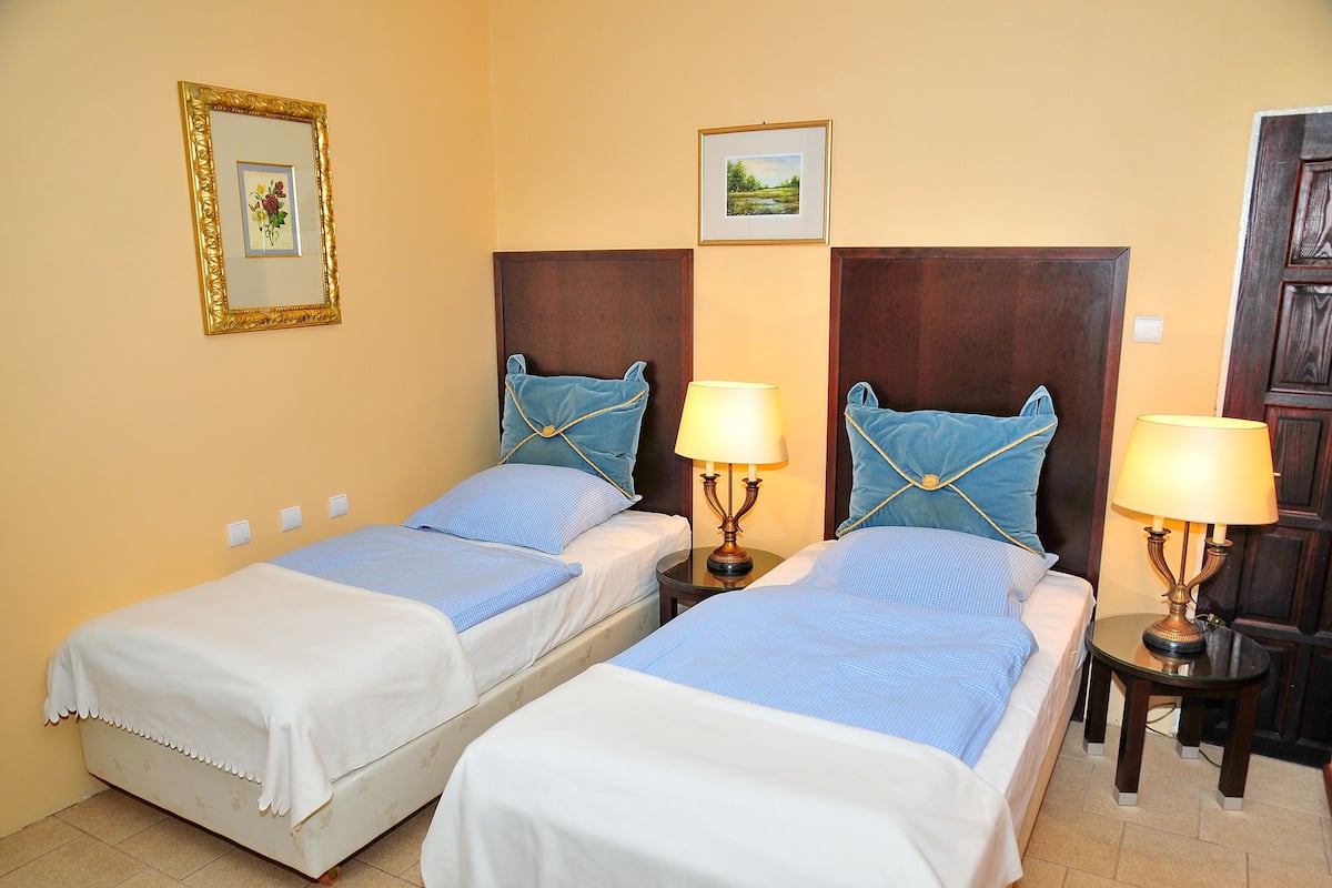 A hotel room with two single beds, each with a blue pillow and a white bedspread. There are two bedside tables with lamps, and the room has a wooden headboard.