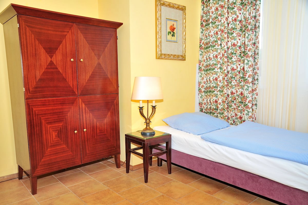 A bedroom with a wooden wardrobe, a bedside table with a lamp, a bed with a blue bedspread, and floral curtains.