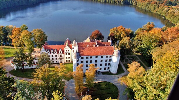 Un grand bâtiment blanc, surmonté d’un toit rouge, entouré d’arbres et bordé par un lac.