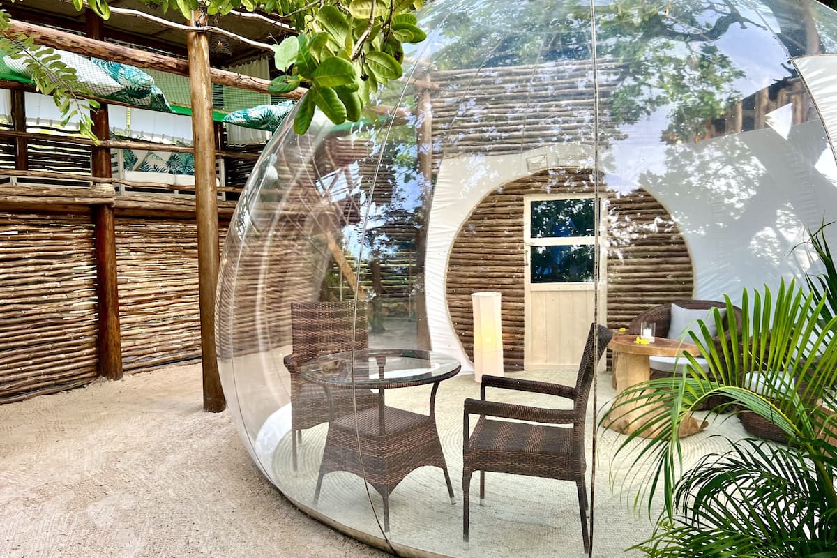 Una estructura en forma de cúpula transparente con mobiliario de mimbre en su interior, que refleja el entorno circundante.