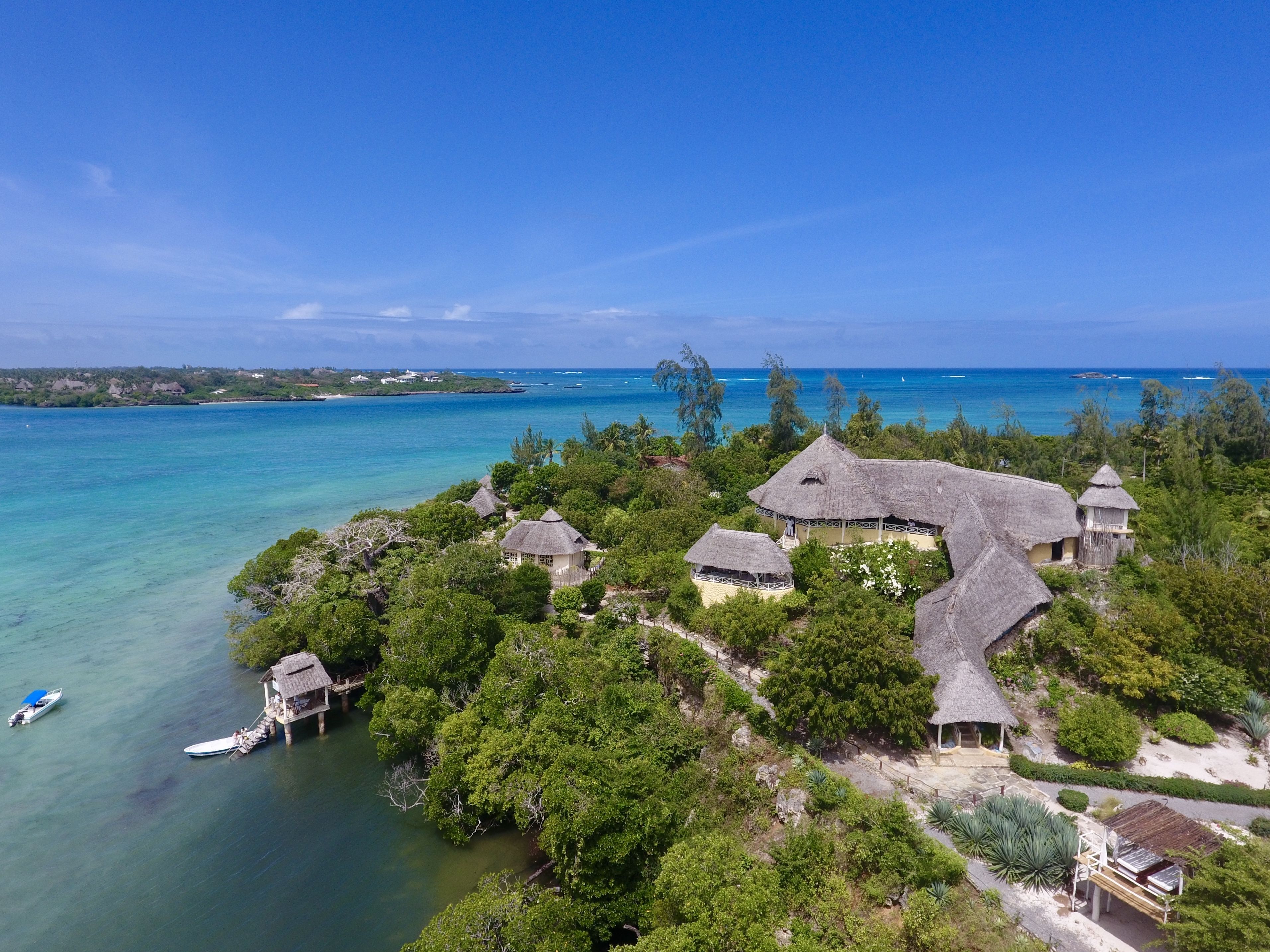 Un complexe hôtelier tropical avec des bâtiments aux toits de chaume, entouré d’une végétation luxuriante et bordé par une mer d’un bleu limpide.